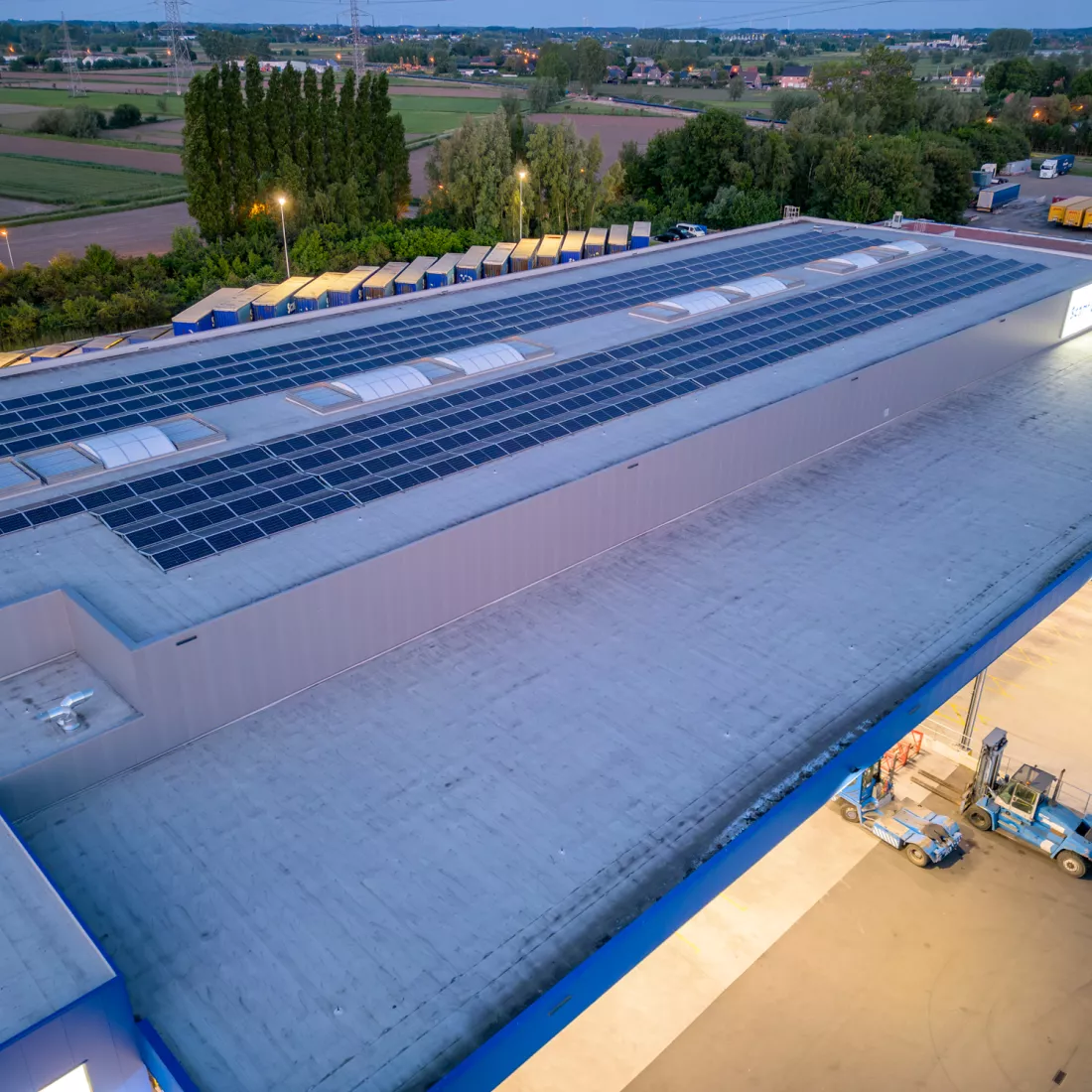 Bovenaanzicht magazijn Scanfor. Het dak is grijs met een blauwe rand. Op het dak liggen zonnepanelen. Achter het magazijn staan containers.