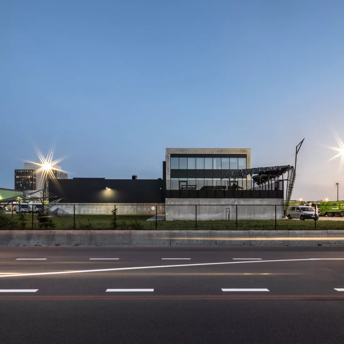 Zijaanzicht sorteer- en recyclagecentrum - Sita kantoor & garage. Links bevindt zich de garage en rechts het kantoor. Het gebouw bestaat uit drie verdiepingen en bevat een balkon. Rond het gebouw is een zwart hek.