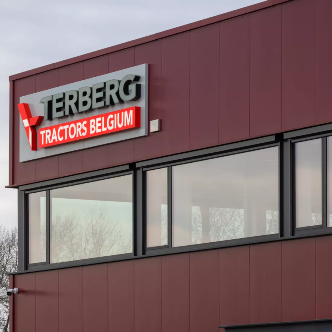 Close-up bedrijfspand Terberg Gent. Het gebouw is rood en heeft ramen met zwarte omkadering. In de linkerhoek van het gebouw hangt het logo van Terberg. Het gebouw is opgebouwd met staal.