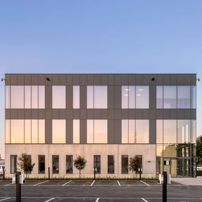 Vooraanzicht kantoor EDR. Het kantoor is opgebouwd met beton. Het gebouw bestaat uit drie verdiepingen. Vanaf de tweede verdieping bestaat het gebouw uit zwarte platen. Het gebouw heeft veel ramen. Rechts bevindt zich de inkom. De inkom bestaat helemaal uit ramen. Voor het gebouw bevindt zich een parking.