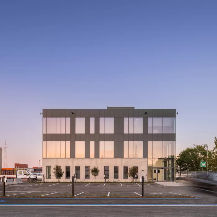 Vooraanzicht kantoor EDR. Het kantoor is opgebouwd met beton. Het gebouw bestaat uit drie verdiepingen. Vanaf de tweede verdieping bestaat het gebouw uit zwarte platen. Het gebouw heeft veel ramen. Rechts bevindt zich de inkom. De inkom bestaat helemaal uit ramen. Voor het gebouw bevindt zich een parking.