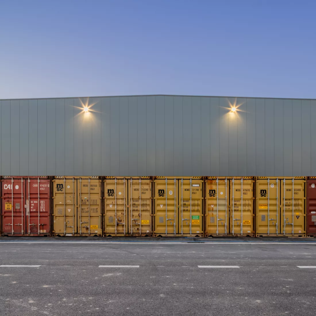 Buitenkant magazijn EDR. Het magazijn bestaat uit grijze smalle platen Voor het magazijn staan rode en gele containers naast elkaar.