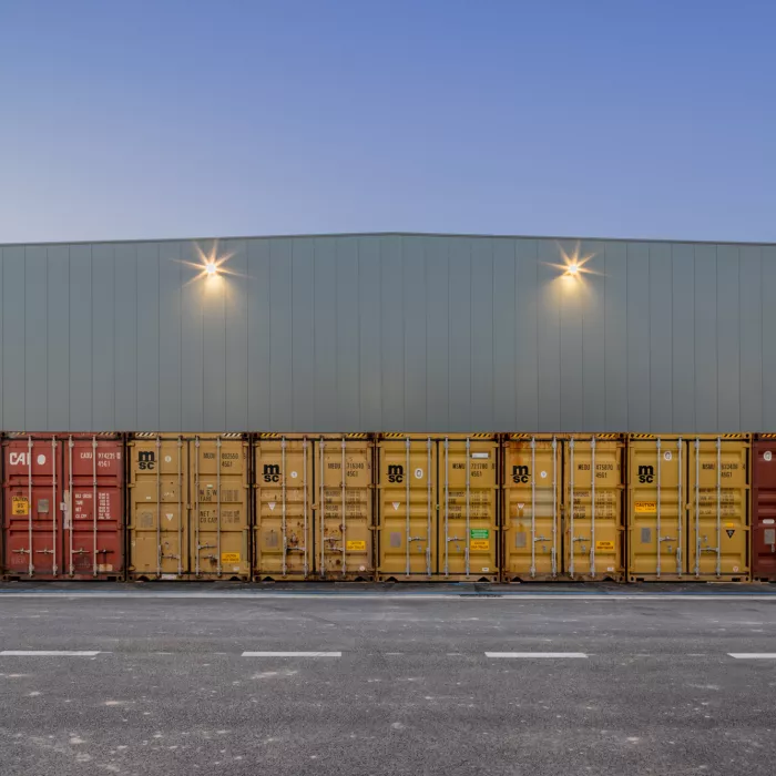 Buitenkant magazijn EDR. Het magazijn bestaat uit grijze smalle platen Voor het magazijn staan rode en gele containers naast elkaar.