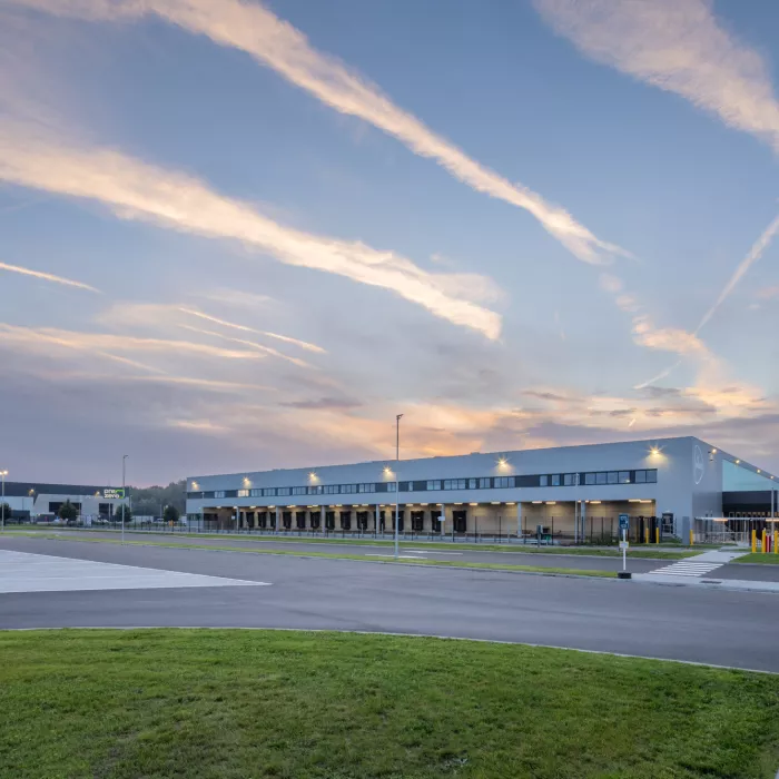 Zijaanzicht sorteercentrum Weerst Evergem (Post NL). Het is gemaakt uit grijs staal. Het bevat een dock voor het laden en lossen van vrachtwagens. Rond het sorteercentrum staat een zwart hek.