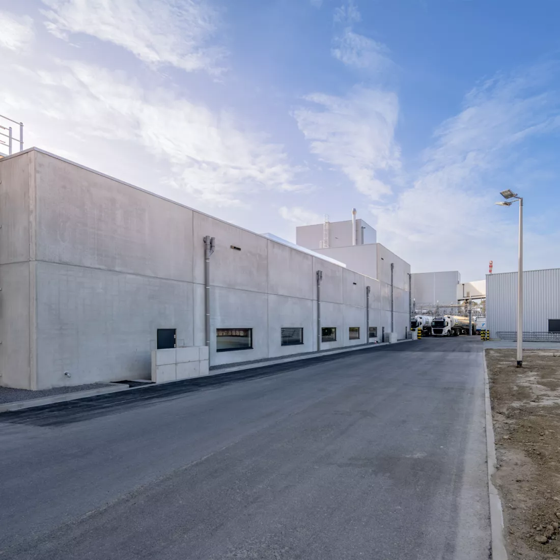Zijaanzicht van de buitenkant van de productiehal van Friesland Campina. De productiehal bevat ook kleine ramen. In de achtergrond staan vrachtwagens.
