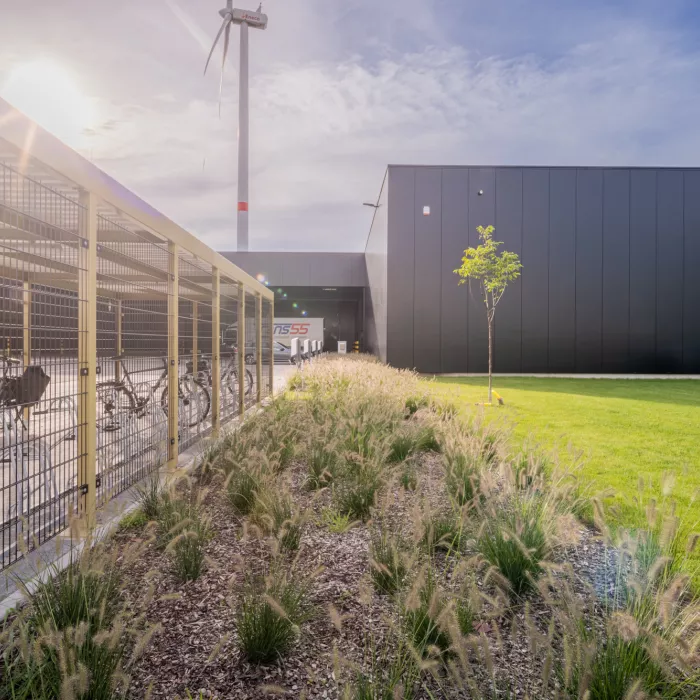 Zijaanzicht magazijn Immo Bolckmans - Proost + mosterdgroene fietsstalling met grasveld en planten ernaast. In de achtergrond staat een witte windmolen.