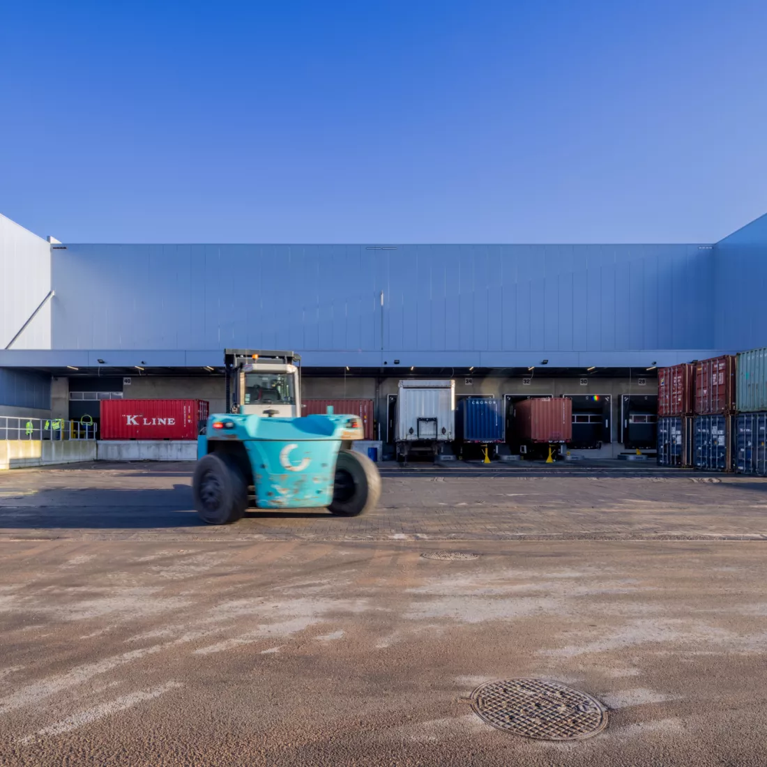 Vooraanzicht dock voor het lossen en laden van vrachtwagens. Voor het dock staan vrachtwagens en containers in verschillende kleuren. De poorten zijn zwart en de muren bestaan uit grijs staal.