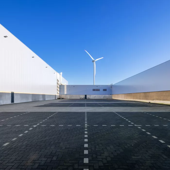 Vooraanzicht kantoor en opslaghal Oostvogels met tussen het gebouw parkingplaatsen. Het gebouw is gemaakt uit grijs staal en bevat kleine zwarte ramen. In de achtergrond staat een witte windmolen.