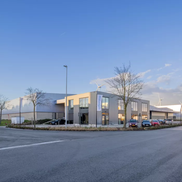 Zijaanzicht kantoor en opslaghal Oostvogels. De foto is gemaakt vanaf de overkant van de straat. De opslaghal bestaat uit grijs staal. Het kantoor bevat grote stalen ramen. Voor het kantoor staan auto's geparkeerd. Ook staan er enkele bomen voor het bedrijf. In de achtergrond zijn witte windmolens te zien.