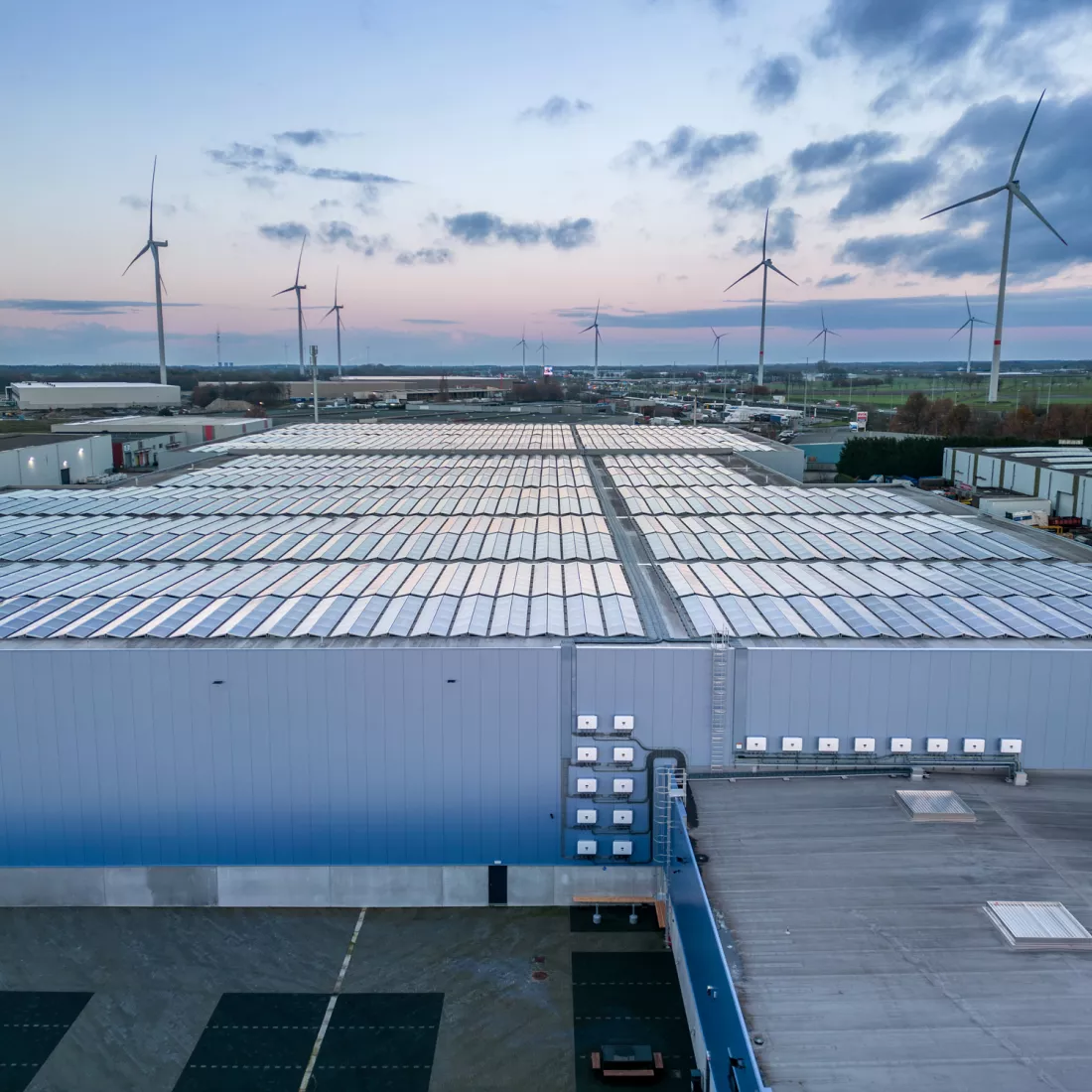 Zijaanzicht opslaghal Oostvogels. Bovenop het dak bevinden zich zonnepanelen. In de achtergrond zijn witte windmolens te zien.