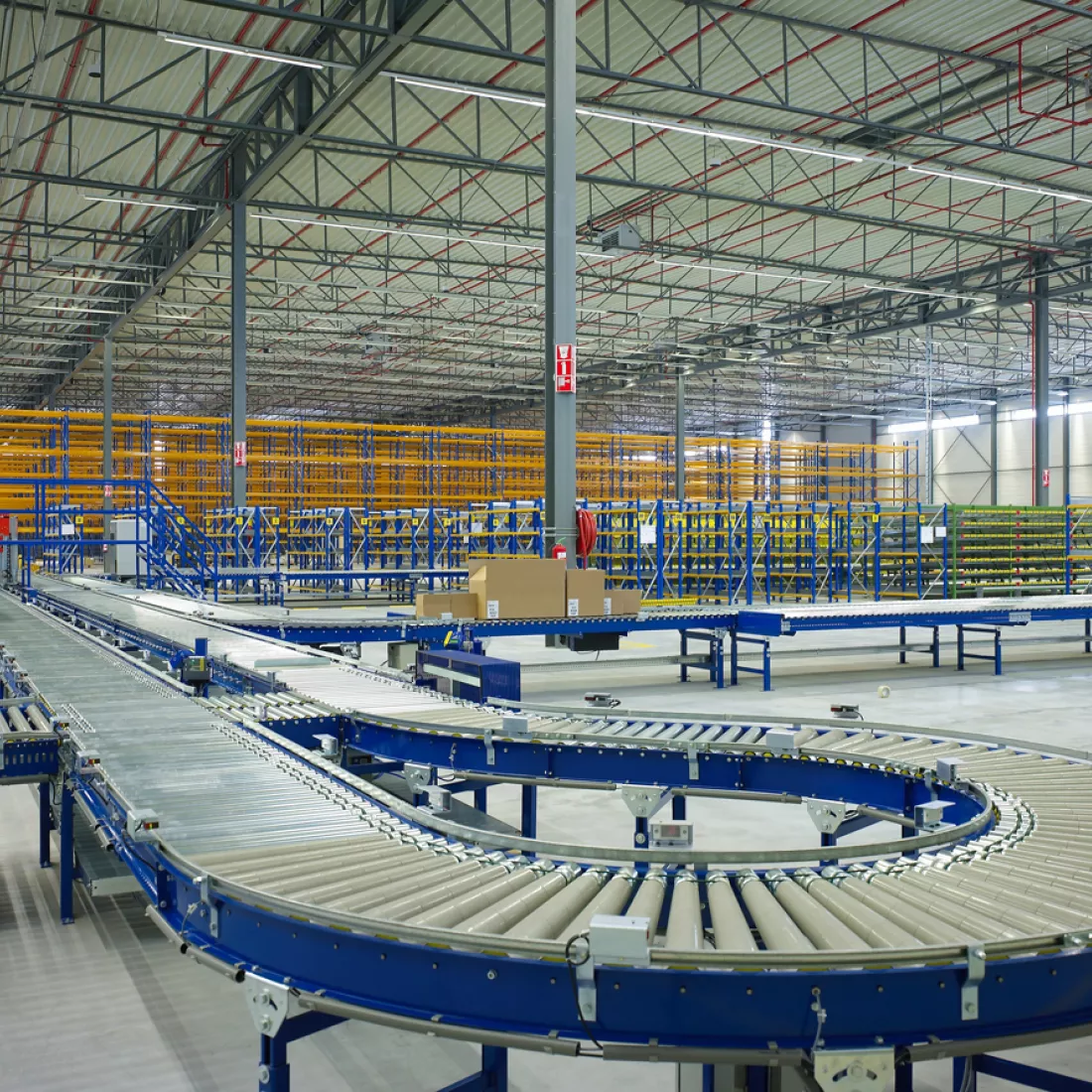 Binnenkant distributiecentrum Prologis met transportband. De transportband is blauw met grijs. Achteraan bevinden zich rekken in het blauw en geel. De hal is gebouwd uit staal.