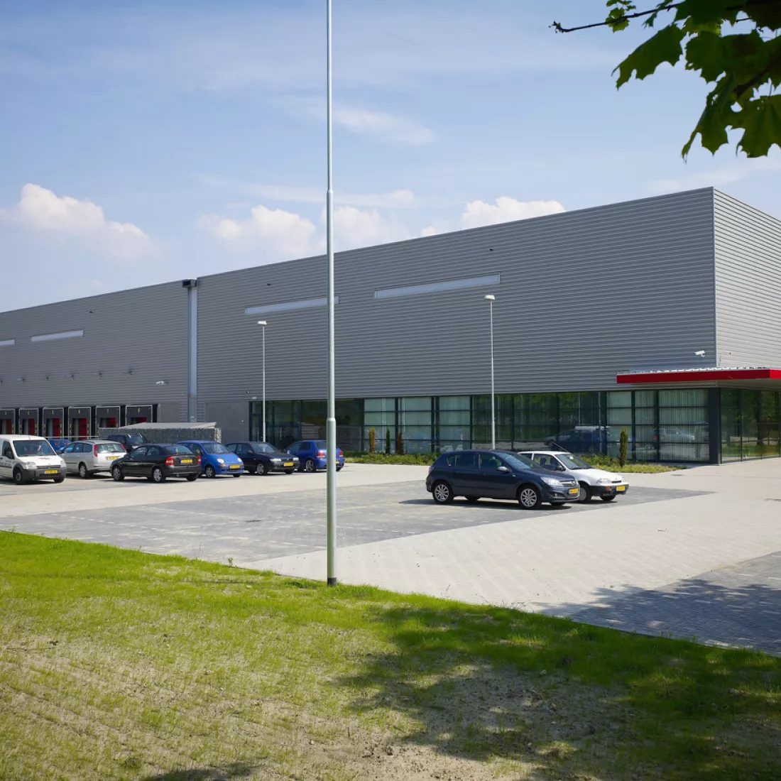 Vooraanzicht distributiecentrum van Prologis Etten Leur.  Het is een een grijs gebouw met rode details en zwarte ramen. Voor het gebouw zijn parkingplaatsen voorzien. Aan de linkerkant van het gebouw bevindt zich een dock voor het laden en lossen van vrachtwagens.
