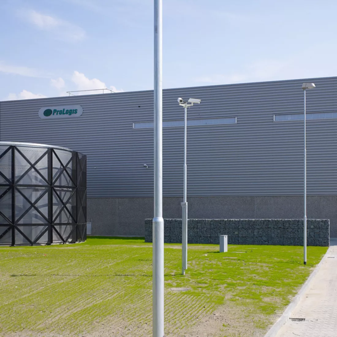 Zijaanzicht Prologis distributiecentrum. Het gebouw is grijs en gebouwd met staal. Links bovenaan bevindt zich het logo van Prologis. Voor het gebouw bevinden zich lantaarnpalen en camerapalen.