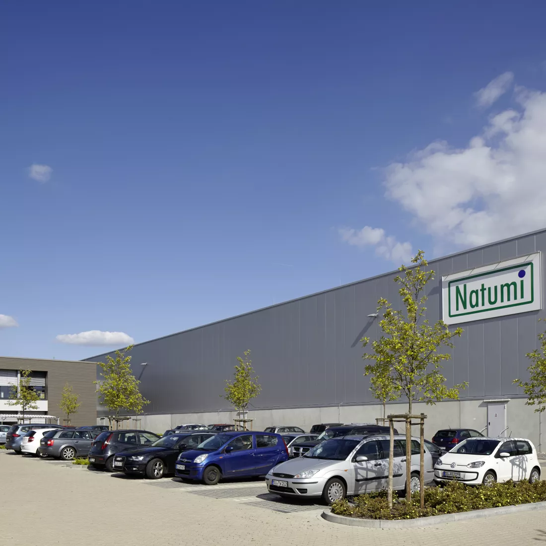 Productiegebouw inclusief kantoren en opslagruimte Natumi Duitsland. Voor het gebouw zijn parkeerplaatsen voorzien. Het gebouw bestaat uit beton en de bovenkant van het magazijn heeft grijze platen. Links bevindt zich het kantoor.