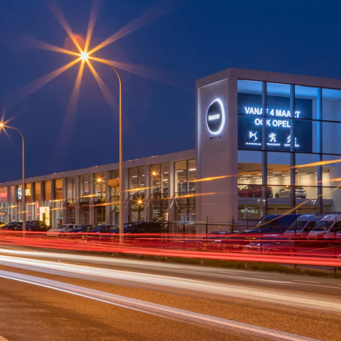 Garage Van Hoecke by night. Het gebouw bestaat uit heel veel ramen. Aan de voorkant hangen verschillende logo's van automerken. Voor het gebouw staan auto's geparkeerd. Het gebouw bestaat uit twee verdiepingen. Op elke verdieping staan auto's tentoongesteld.