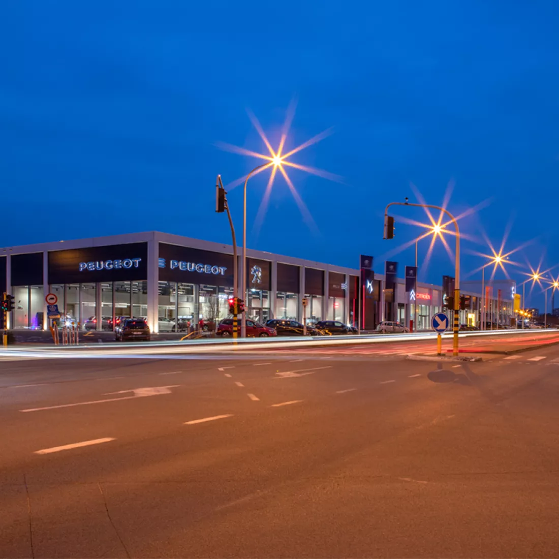 Zijaanzicht Garage Van Hoecke by night. Het gebouw bestaat uit een groot deel uit ramen. Bovenaan hangen verschillende logo's van automerken.