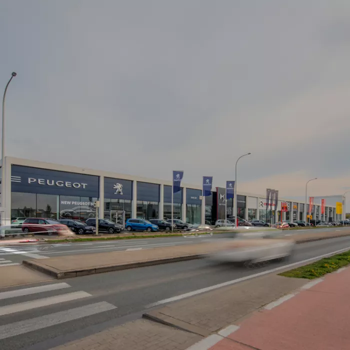 Vooraanzicht garage Van Hoecke. Het gebouw is opgebouwd met een combinatie van materialen. Het bevat hele grote ramen. Bovenaan hangen verschillende logo's van automerken. Bij elk logo staan er ook vlaggen van dit merk voor het gebouw. Elk merk heeft een andere kleur van platen tegen de muur hangen. Voor het gebouw staan auto's geparkeerd.