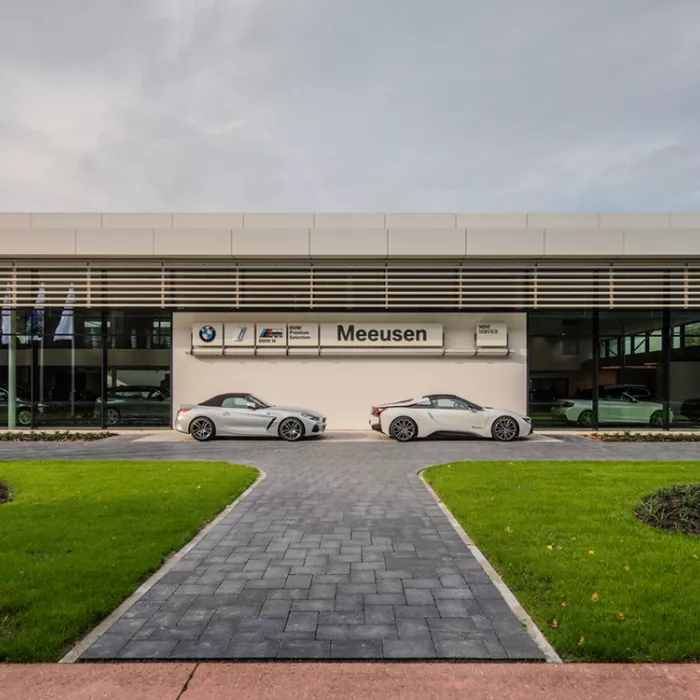 Close-up vooraanzicht showroom garage Meeusen. Er loopt een pad naar de garage. Langs het pad zijn grasvelden. Het midden van het gebouw bevat een witte muur met het logo erop. Aan de linker- en rechterkant zijn grote ramen om de showroom te zien. Ervoor staan twee auto's.