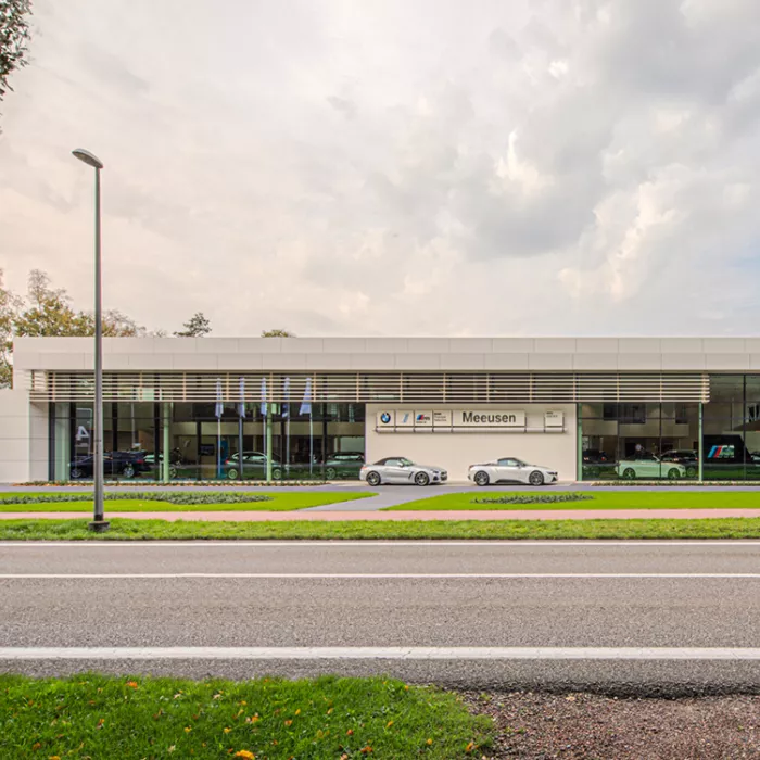 Vooraanzicht showroom garage Meeusen. Er loopt een pad naar de garage. Langs het pad zijn grasvelden. Het midden van het gebouw bevat een witte muur met het logo erop. Aan de linker- en rechterkant zijn grote ramen om de showroom te zien. Ervoor staan twee auto's.