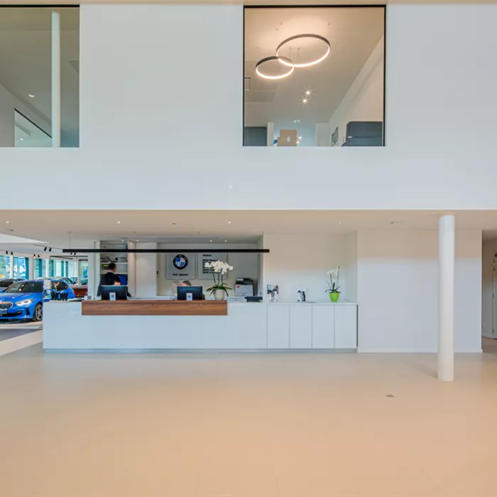 Binnenkant showroom garage Meeusen. De muren zijn wit. Boven de showroom bevindt zich het kantoor. Links staan auto's ten toon gesteld. Rechts is een onthaalbalie met twee computers.