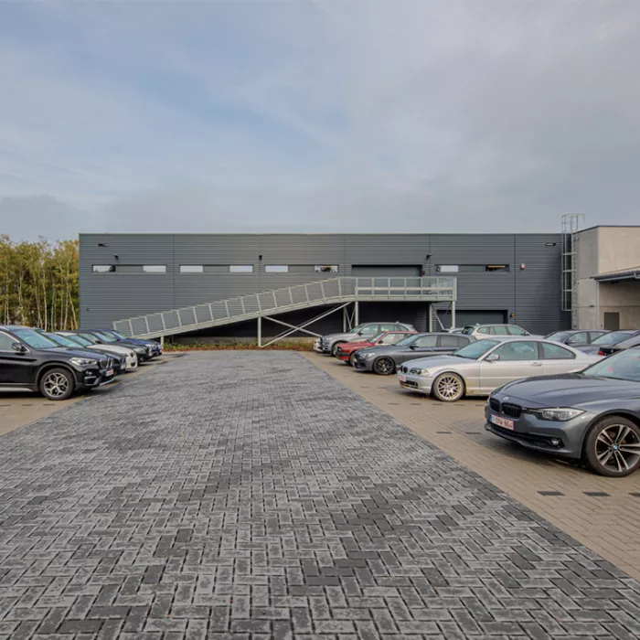 Achterkant showroom en werkplaats garage Meeusen. Het gebouw is donkergrijs en bevat zwarte smalle ramen. Voor het gebouw staan auto's geparkeerd op parkeerplaatsen.