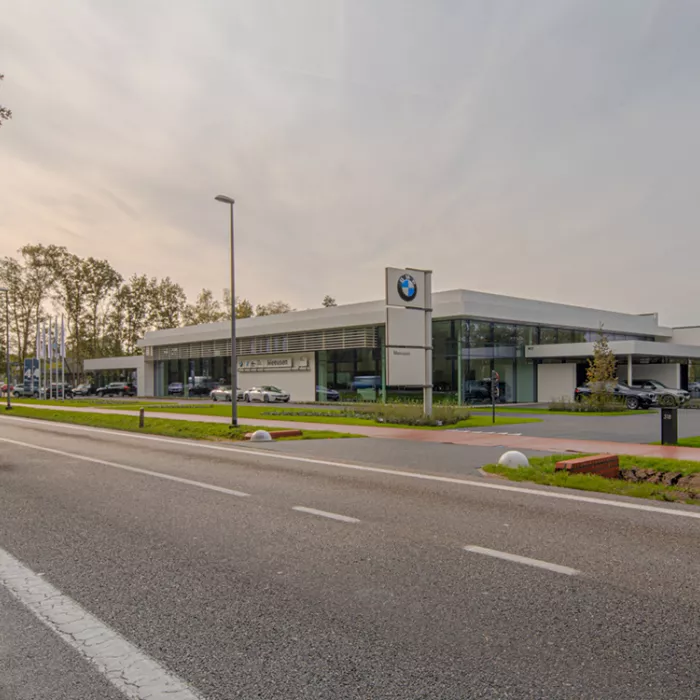 Zijaanzicht showroom garage Meeusen. De afbeelding is gemaakt vanaf de overkant van de straat. Rechts naast het gebouw zijn parkeerplaatsen voorzien. Het is een wit gebouw met grote ramen. Voor het gebouw staat een paal met drie borden. Op het bovenste bord hangt het BMW logo en op het onderste staat Meeusen.