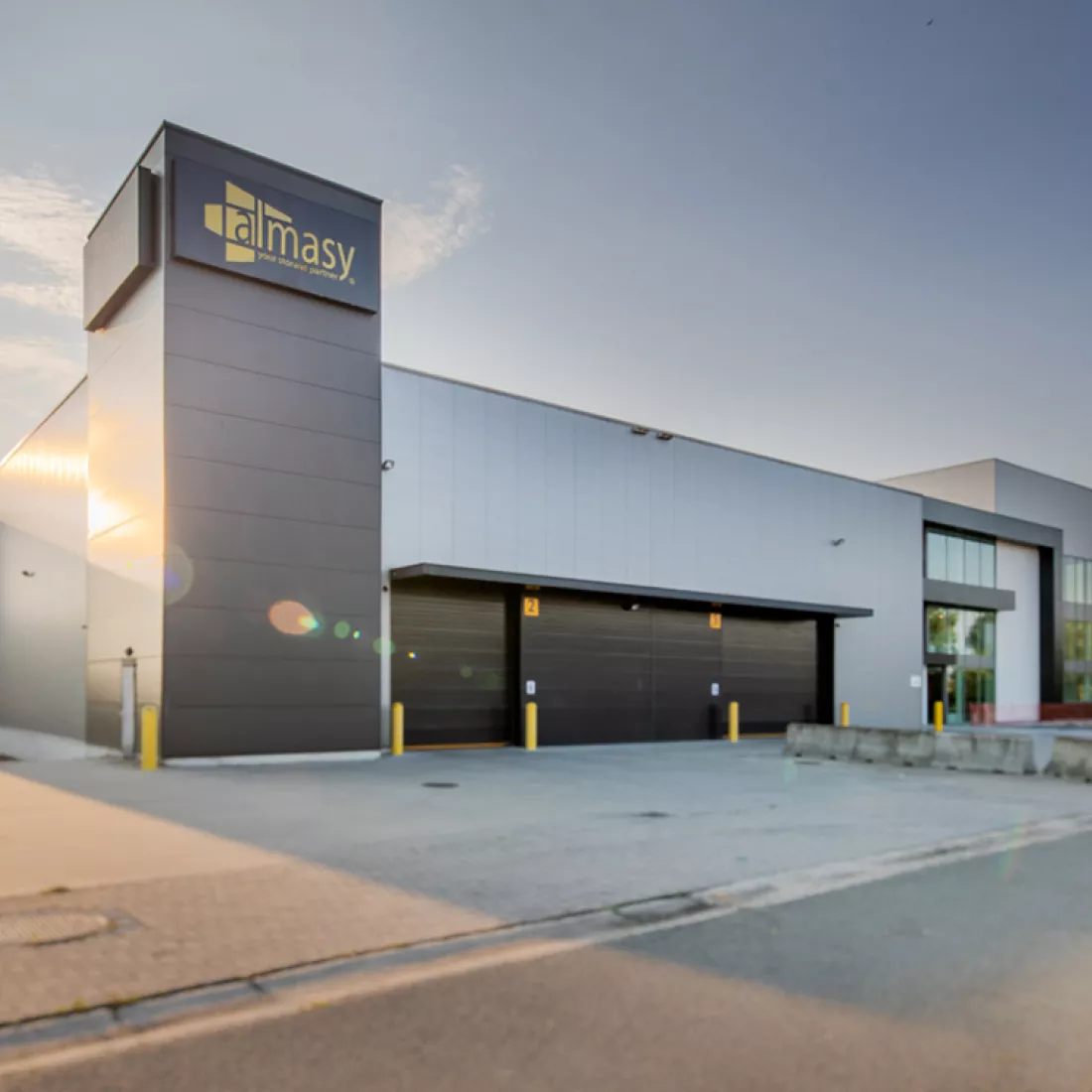 Zijaanzicht Almasy kantoor. Links bevindt zich het bestaande industriegebouw en rechts het kantoor dat bijgebouwd is. Het gebouw bestaat uit grijze en zwarte platen. Links hangt het logo van Almasy. Het industriegebouw bevat zwarte poorten. Het nieuwe kantoor bevat veel ramen.