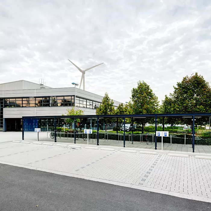 Parking voor het distributiecentrum van Donaldson. Er is een parking voor auto's voorzien en daarachter een parking voor fietsers. Links achter de fietsparking bevindt zich het distributiecentrum. Rechts daarvan staan bomen.