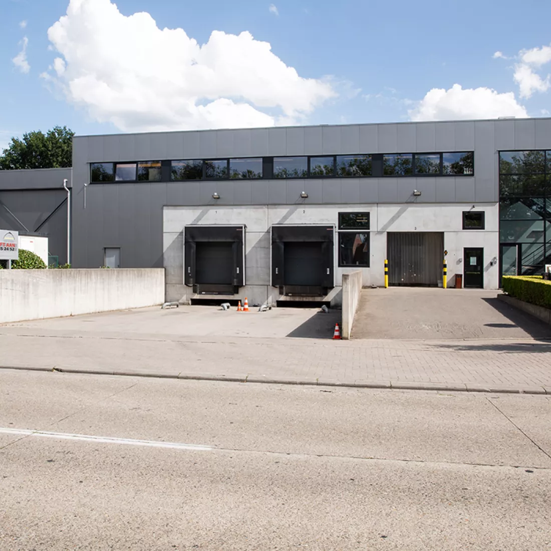 Vooraanzicht werkgebouw Kelkro. Het gebouw bestaat uit twee verdiepingen. Het gebouw bestaat uit beton en grijze platen. Ook bevat het enkele ramen met zwarte omrandig. Er is een dock voor het laden en lossen van vrachtwagens. Het dock bevat zwarte poorten.