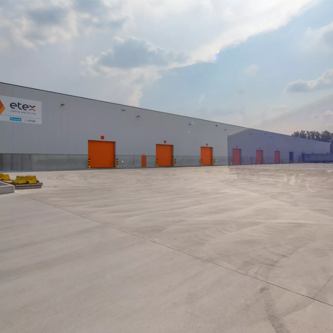 Zijaanzicht magazijn Promat hal 9. Het gebouw is grijs en heeft oranje poorten. Tijdens de opbouw is staal gebruikt. Het logo van Etex hangt links.