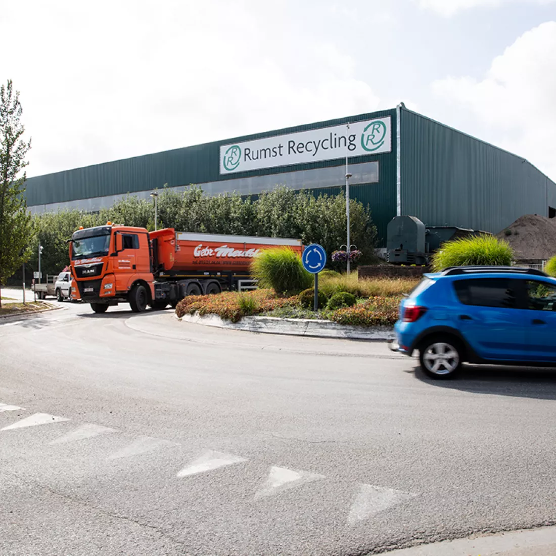 Zijaanzicht grondhal Rumst Recycling. Het gebouw bestaat uit donkergroene platen. Rechts bovenaan hangt het logo. Voor het gebouw bevindt zich een straat met een rondpunt.