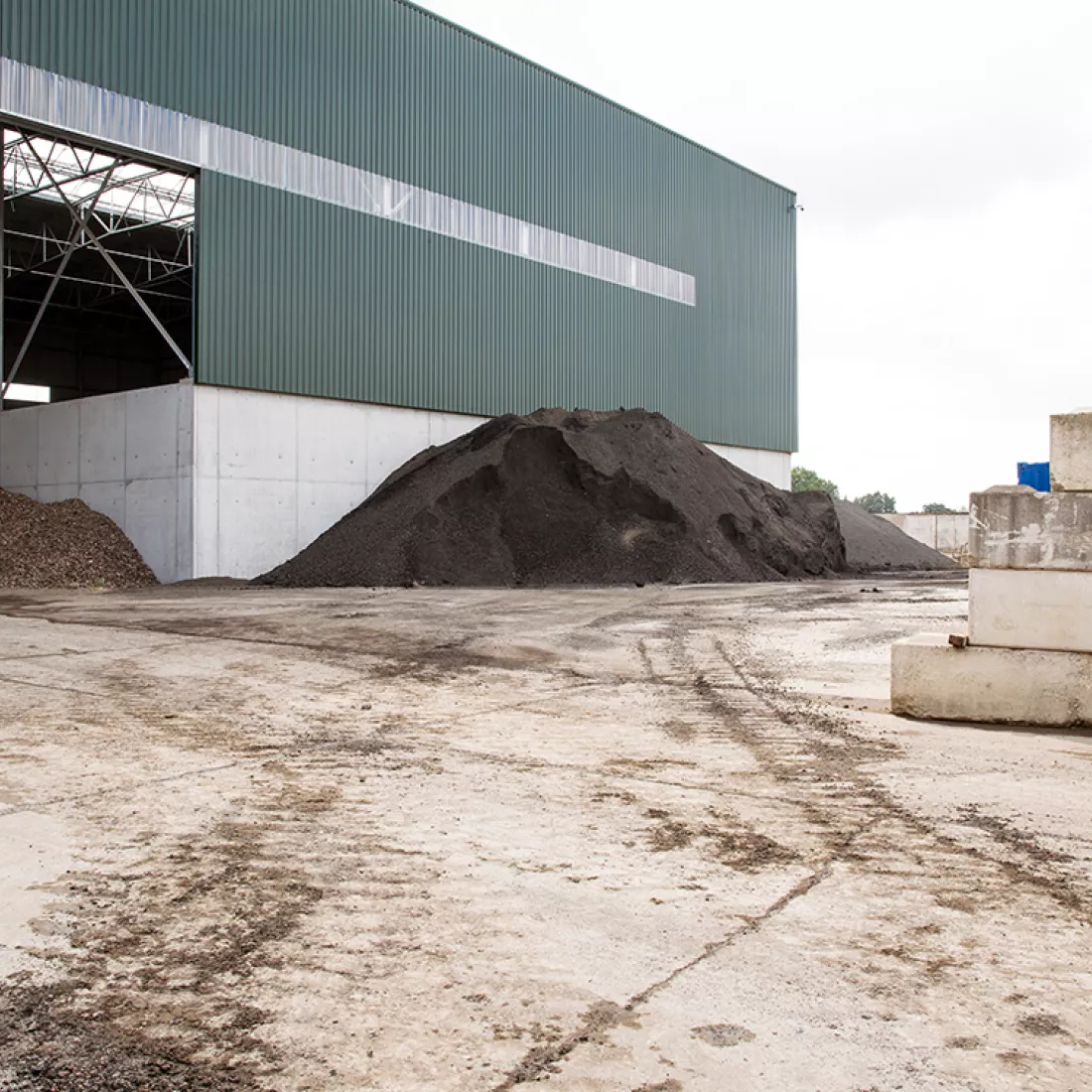 Zijaanzicht grondhal Rumst Recycling. Deze zijaanzicht afbeelding is gemakt vanaf het bedrijfsterrein. Links bevindt zich de hal. Voor de hal ligt een berg zand.