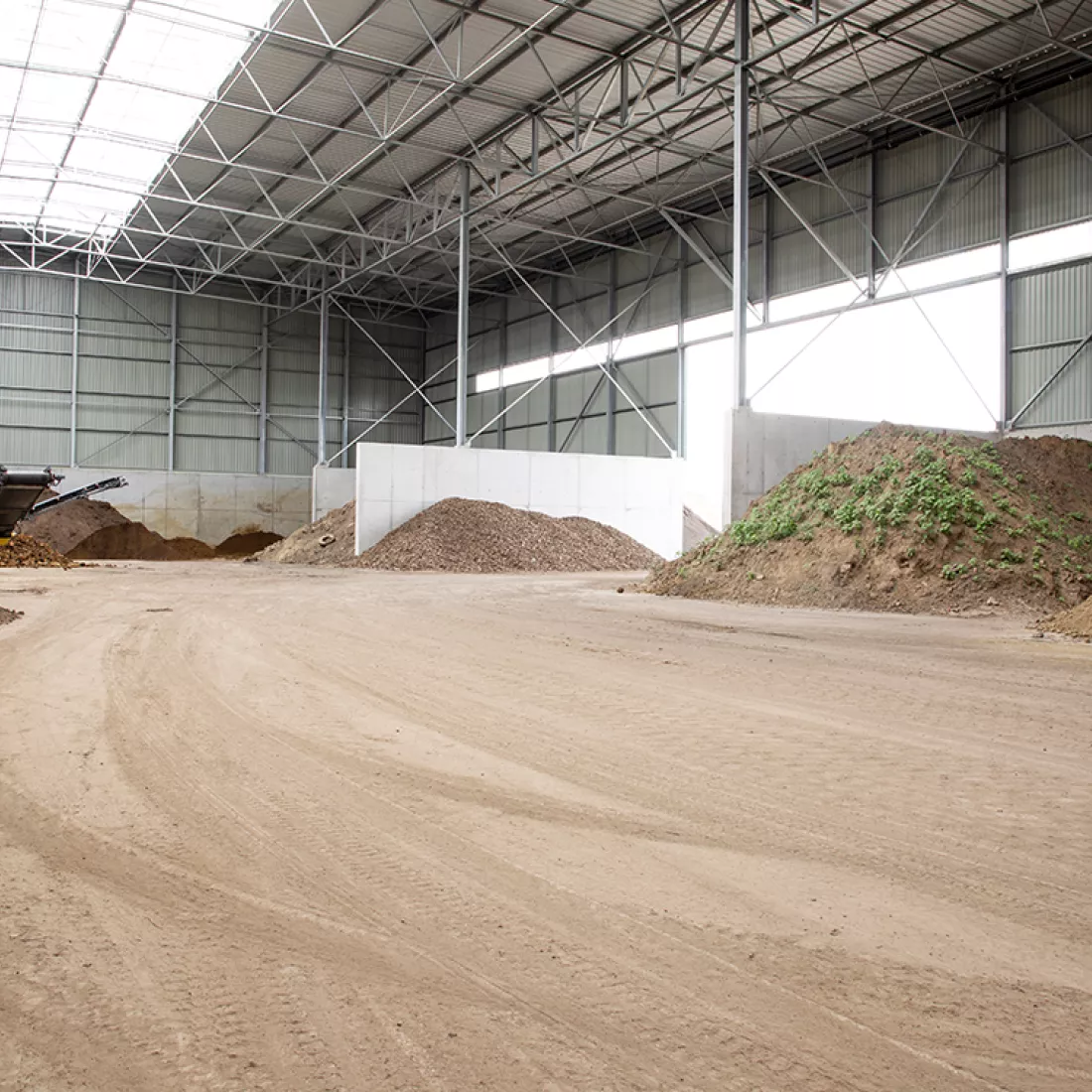 Binnenkant grondhal Rumst Recycling. Rechts heeft de hal een grote opening in de muur. In het plafond bevindt zich een grote lichtstraat. Het zand is verdeeld door betonnen muren.