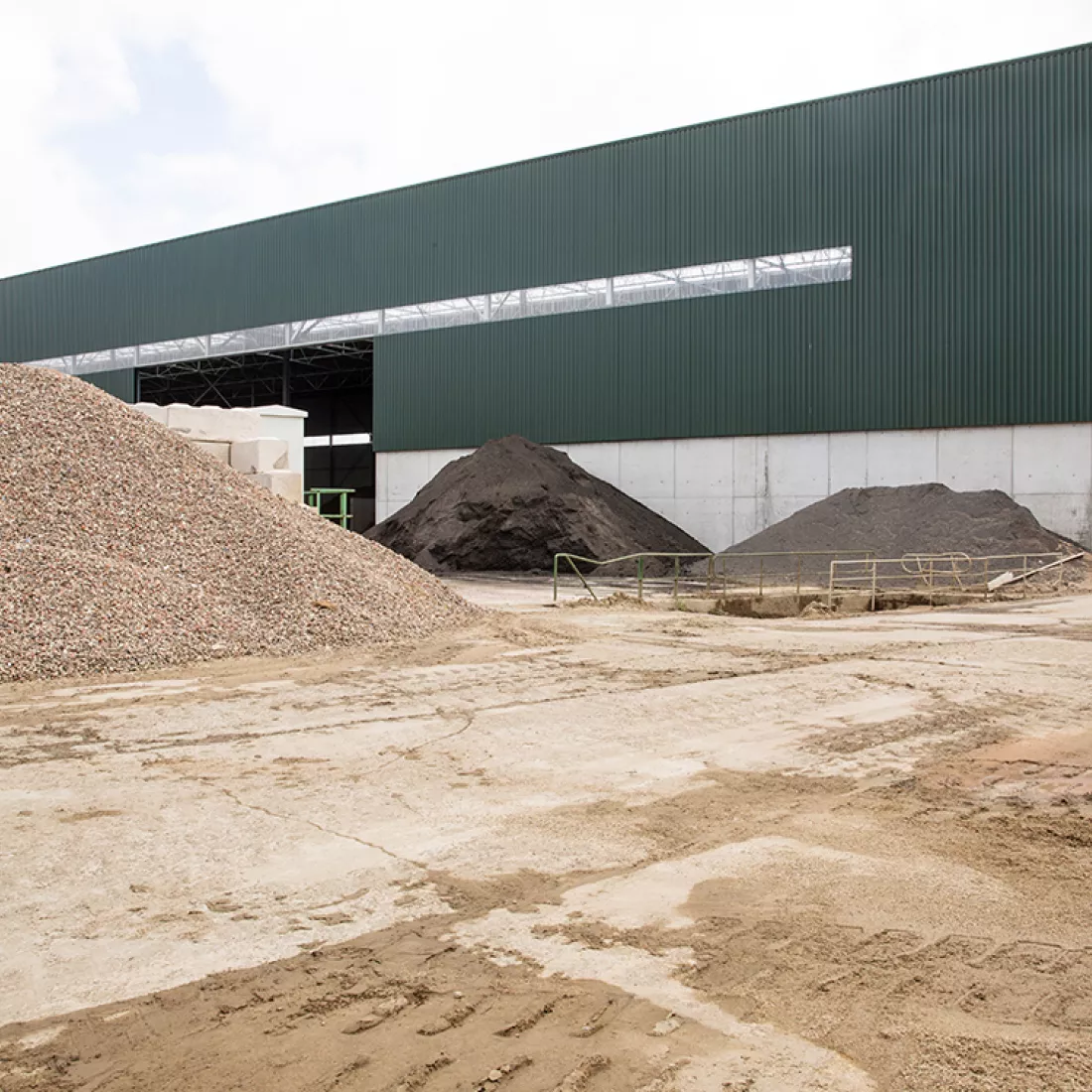 Zijaanzicht grondhal Rumst Recycling. Rond het gebouw liggen verschillende bergen zand.