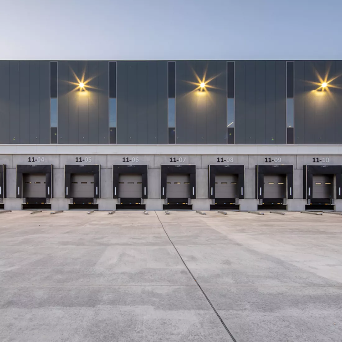 Vooraanzicht dock - DC Braak. Hier laden en lossen vrachtwagens. Het dock bevat grijze poorten. Er rond is een betonlook. Boven het dock bestaat uit het gebouw uit donkergrijze platen en een paar smalle ramen.