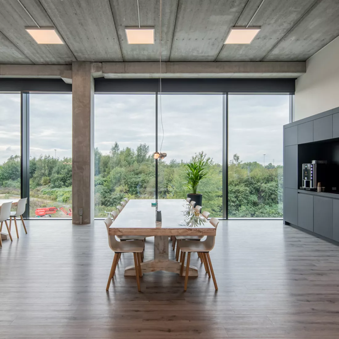 Binnenkant kantoor De Wille Fase 8. De afbeelding is van de lunchruimte. Rechts staat een zwart keukenblok met koffiemachine op. In het midden staat een lange rechthoekige tafel met witte stoelen rond. De muur achter de tafel heeft grote ramen.
