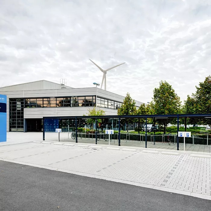 Zijaanzicht distributiecentrum Donaldson. Het is gebouwd met staal en hout. Het is grijs gebouw. Voor het gebouw is parking voorzien voor auto's en fietsen.