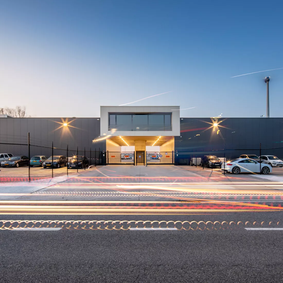 Vooraanzicht werkgebouw Thys Schenk. Het gebouw is zwart en de tweede verdieping in het midden wit. Rond het gebouw is een zwarte omheining. Links hangt het logo Schenk en rechts Thys.