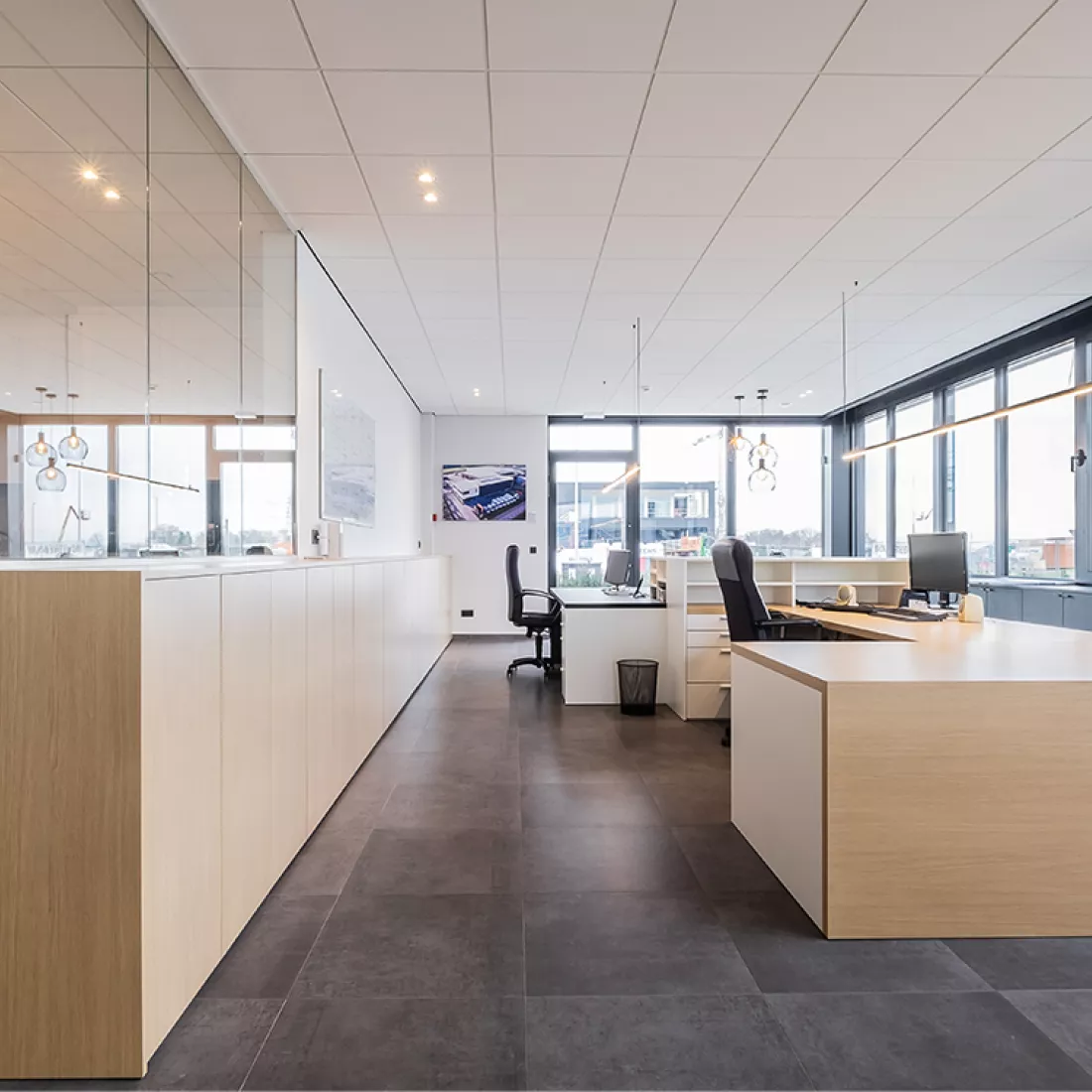 Binnenkant kantoor Van Den Bogerd. De bureau en kasten zijn uit hout. De vloer is een zwarte steen en de muren zijn wit. Links staan de kasten en rechts de bureaus. Tegen de linker muur hangt ook een spiegel.