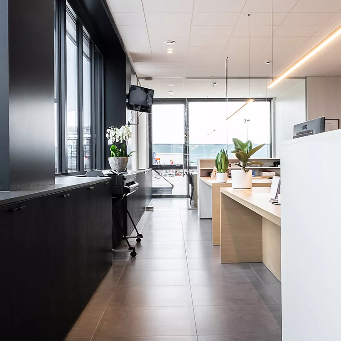 Binnenkant kantoor Van Den Bogerd. Links staan zwarte kasten. De muur bestaat uit ramen met zwarte omkadering. Rechts staan de bureaus gemaakt uit hout.