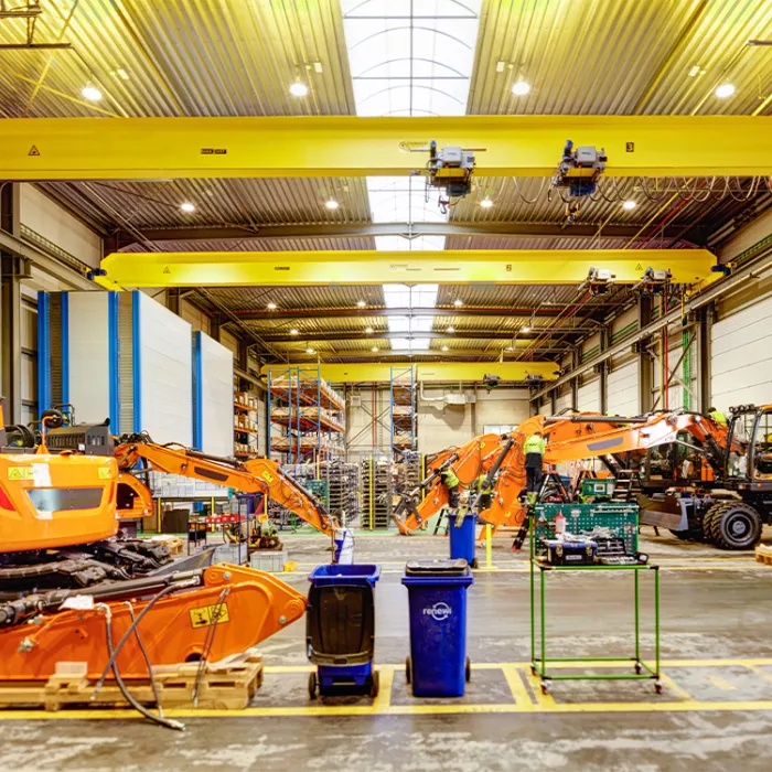 Binnenkant werkplaats Wallenius. Het gebouw is gebouwd met staal. Bovenaan hangen gele balken. In het midden van het dak is een lange glazen koepel. Er staan verschillende onderdelen van werkmachines. Alles is op de grond afgebakend met gele lijnen. Links vanachter staan rekken met materiaal in gestockeerd.