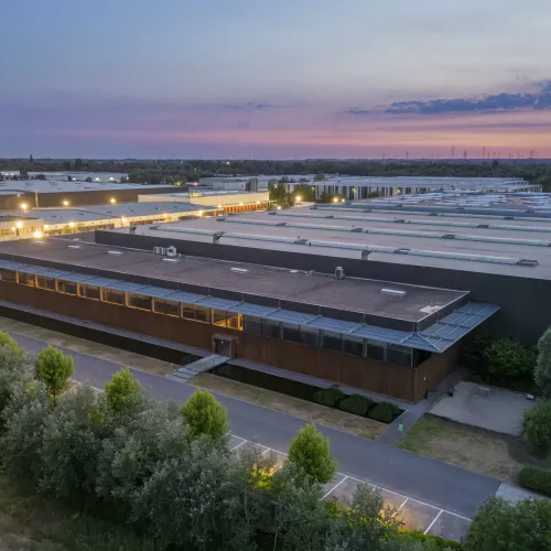 Een luchtfoto van een moderne industriële faciliteit tijdens de schemering. Het uitgestrekte gebouw met plat dak heeft talrijke dakramen en luchtunits, en is omgeven door weelderig groen en een goed onderhouden landschap. De gevel van het gebouw is deels bekleed met hout, waardoor het geïntegreerd wordt met de natuurlijke omgeving. De lucht is geschilderd met zachte tinten paars en blauw, wat de overgang van zonsondergang naar avond suggereert.