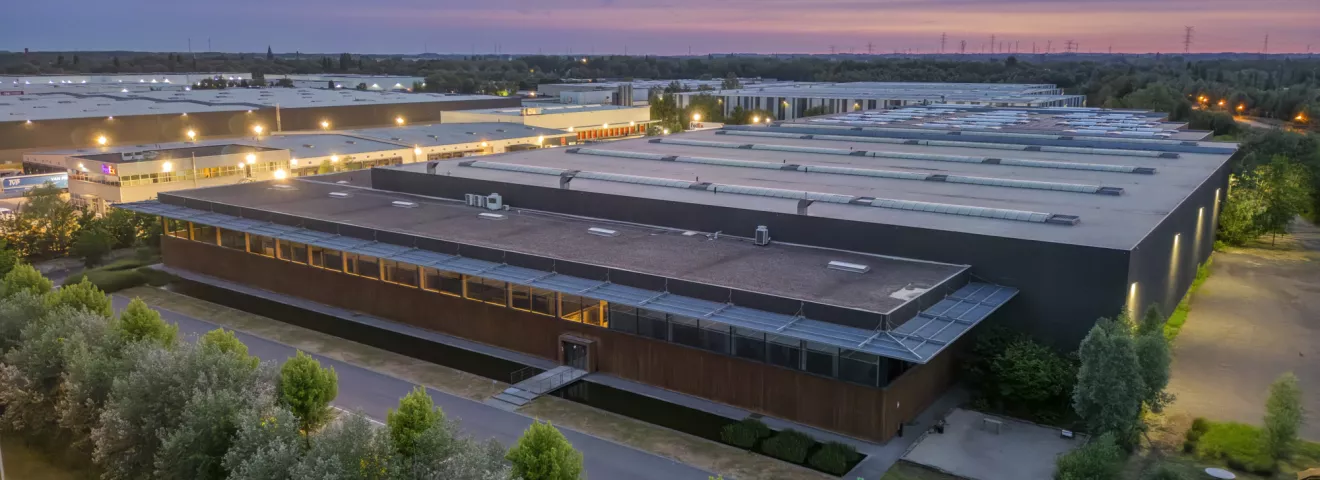 Een luchtfoto van een moderne industriële faciliteit tijdens de schemering. Het uitgestrekte gebouw met plat dak heeft talrijke dakramen en luchtunits, en is omgeven door weelderig groen en een goed onderhouden landschap. De gevel van het gebouw is deels bekleed met hout, waardoor het geïntegreerd wordt met de natuurlijke omgeving. De lucht is geschilderd met zachte tinten paars en blauw, wat de overgang van zonsondergang naar avond suggereert.