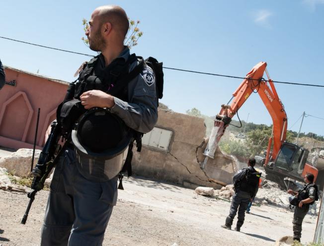 Een Israelische soldaat met een oorlogswapen staat voor een bulldozer die een huis aan het vernietigen is.