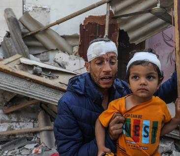 Foto van een gewonde Palestijne man met een kind, met een vernield huis op de achtergrond