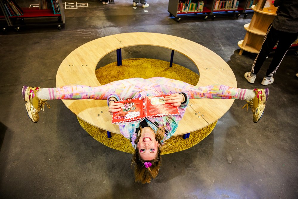 Kleurrijk gekleerde jonge dame ligt in spagaat op een ronde tafel terwijl ze een boek leest