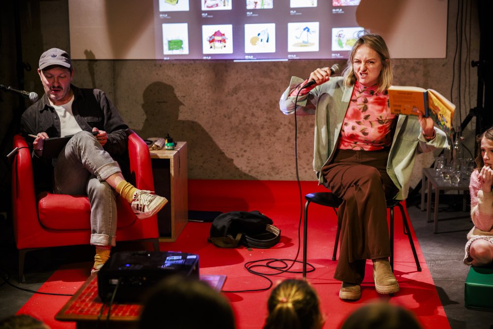 Auteur trekt een gek gezicht tijdens voorlezen van een boek, naast haar zit een tekenaar te schetsen