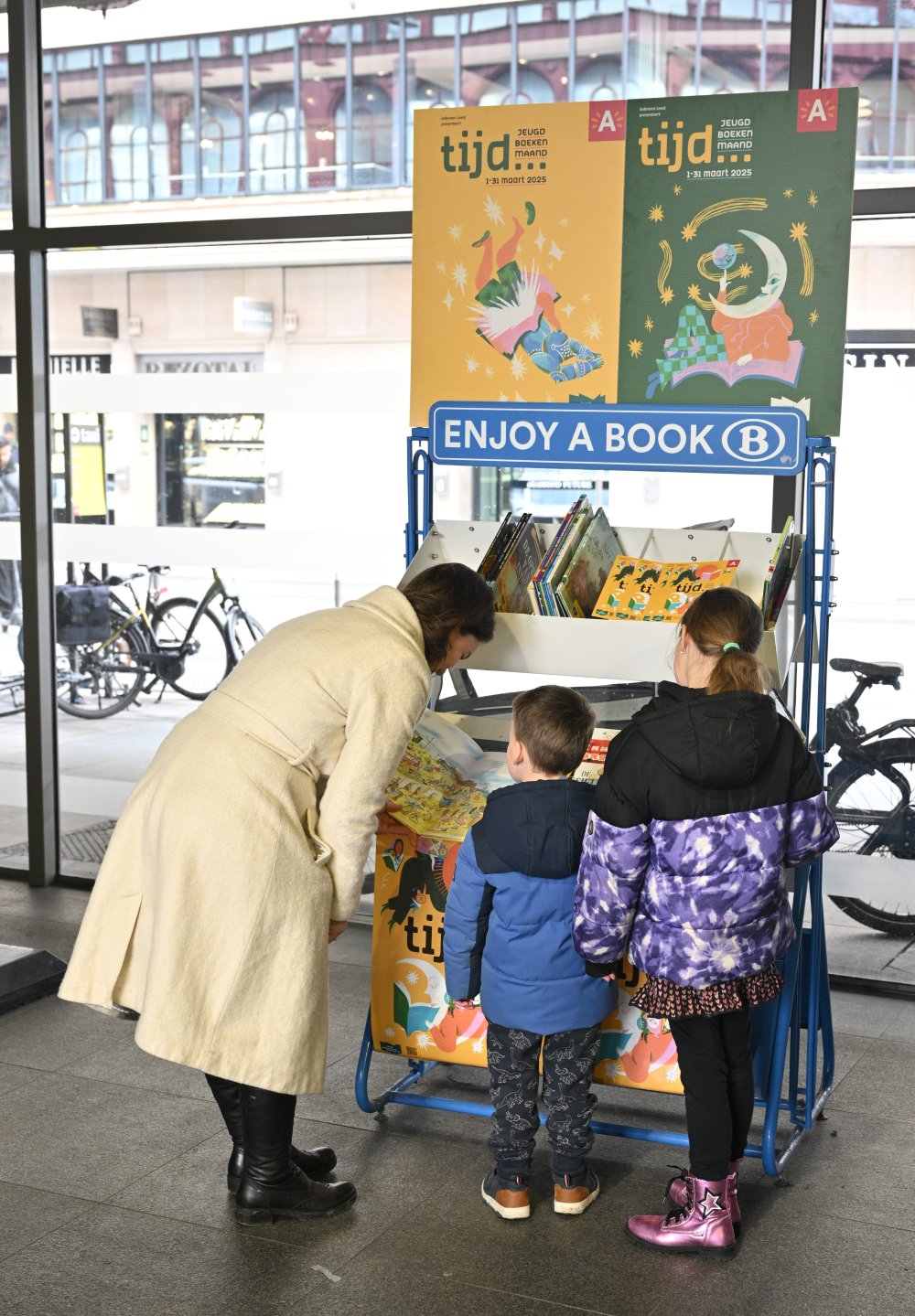 Een mama staat met haar twee kinderen een boek uit te kiezen bij een boekenruilkast van de NMBS aangekleed in het thema Tijd