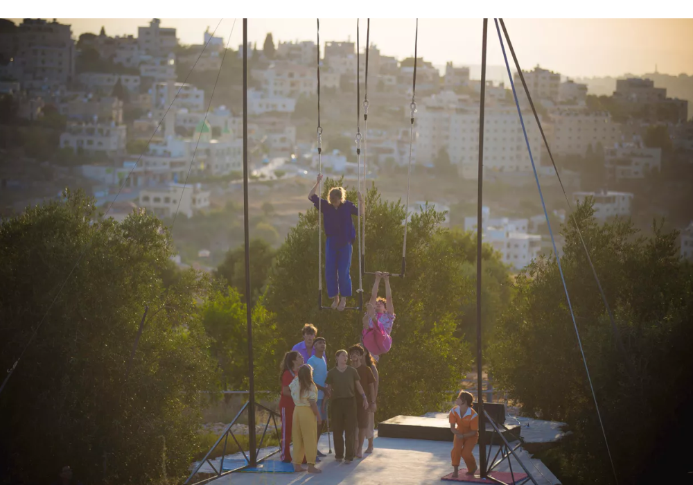 Een groep jonge circusartiesten staat op een verhoogd platform buiten met een stad op de achtergrond. Twee mensen hangen aan trapezes boven het platform, terwijl anderen toekijken. De zon schijnt helder.