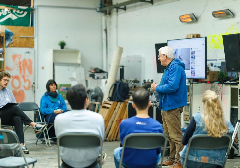 Ton geeft een presentatie aan een kleine groep mensen die in een halve cirkel zit, in een informele ruimte met industrieel karakter.
